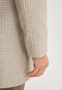 Lichtbeige jas met een geruit patroon, voorzien van een gladde textuur en een getailleerde pasvorm. Close-up van de mouw en hand zichtbaar.