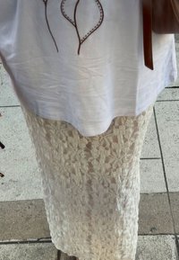 Hvid top med broderet bladdesign over en gennemsigtig blondekjole med blomstermønster. Brun taske holdes i venstre hånd.