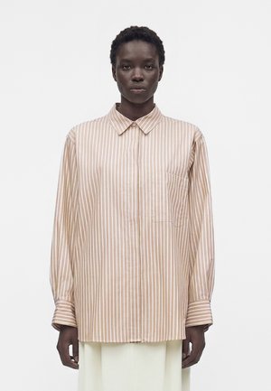 Sötét bőrszínű felnőtt, bézs-fehér függőleges csíkozású, galléros, hosszú ujjú ingben, fehér egyszínű háttér előtt áll.