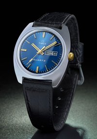 Montre à quartz Timex avec un cadran bleu, des aiguilles dorées et un affichage de la date. Elle dispose d'un boîtier argenté et d'un bracelet en cuir noir avec une finition texturée.