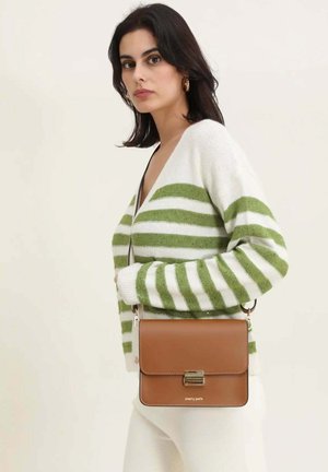 Femme portant un pull rayé blanc et vert tenant un sac bandoulière en cuir marron avec une boucle dorée.