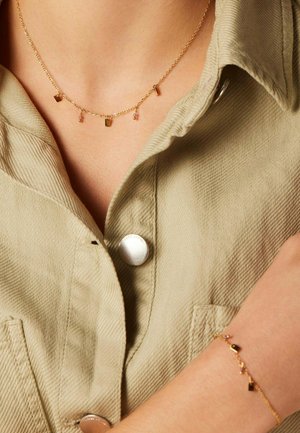 Collier doré et bracelet assorti avec de petits pendentifs géométriques et des accents pastel. Porté sur une chemise beige texturée avec un détail de bouton.