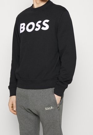 Person iført en sort sweatshirt med teksten "BOSS" og grå joggingbukser med "BALR." logo, hånd i lommen.