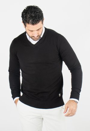 Pull léger noir en v avec manches longues et poignets côtelés, superposé à une chemise à col blanc, comprenant un petit logo en bas.