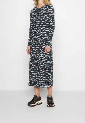 Kobieta ubrana w czarno-białą sukienkę w nadruk zebry o długości sięgającej do połowy łydki i z długim rękawem, nosząca masywne czarne sneakersy, na jednolitym białym tle.