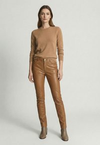 Vrouw met een camelkleurige trui, bruine leren broek en grijze enkellaarzen, staand tegen een effen lichtgrijze achtergrond.