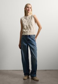 Junge Frau mit langen blonden Haaren, die einen cremefarbenen ärmellosen Zopfmuster-Pullover, weit geschnittene blaue Jeans und beige Sneakers trägt, vor einer schlichten Wand.