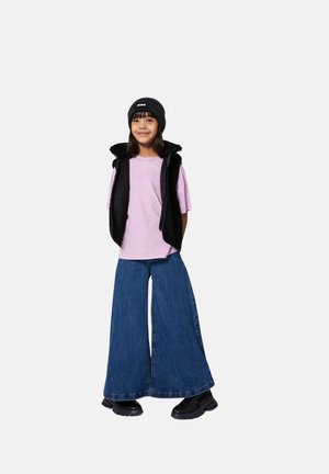 Kind draagt een zwarte beanie, een zwarte mouwloze jas, een lichtpaarse T-shirt, wijde blauwe spijkerbroek en zwarte laarzen, staat rechtop en glimlacht.