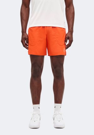 Hombre que lleva puestos unos pantalones cortos deportivos naranja brillante, calcetines blancos, zapatillas blancas Nike y una camiseta sin mangas blanca, de pie mirando hacia adelante.