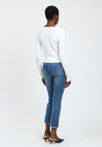 Top blanco ajustado de manga larga con cuello redondo, combinado con vaqueros recortados azules con dobladillo deshilachado y bolsillos traseros. Zuecos negros de tacón.