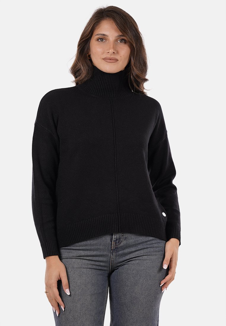 Donna che indossa un maglione nero a collo alto e jeans grigi, in piedi contro uno sfondo bianco semplice, rivolta in avanti.