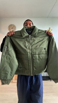 Olivno zelena bomber jakna z zadrgo spredaj, dvema velikima sprednjima žepoma in stranskim žepom. Mehka tekstura in oblikovan ovratnik.