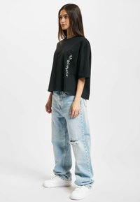 Schwarzes, kurzes T-Shirt mit weißem Grafikdruck auf der Vorderseite, kombiniert mit hellblauen, ausgewaschenen, lässigen Jeans und weißen Sneakern.