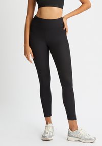 Leggings negras de canalé con una cintura alta, que presentan una textura suave. Combinados con zapatillas de correr gris claro, que exhiben un diseño elegante.
