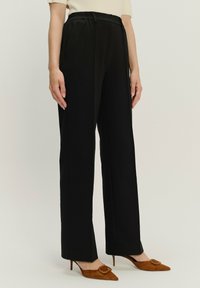 Schwarze, hochgeschnittene Hose mit lockerer Passform, ausgestattet mit seitlichen Taschen und einer glatten Textur, kombiniert mit spitzen, braunen Wildlederschuhen.
