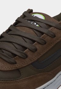 Κοντινό πλάνο καφέ σουέντ αθλητικού παπουτσιού με σκούρα κορδόνια, σήμα Nike στο πλάι και κυκλικό λογότυπο στη γλώσσα με πράσινη λεπτομέρεια.