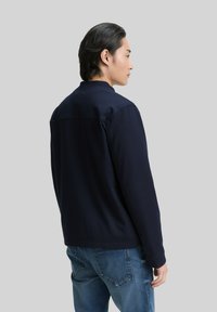 Man met zwart haar, gekleed in een donkerblauwe jas en blauwe spijkerbroek, die zijwaarts staat tegen een effen grijze achtergrond.