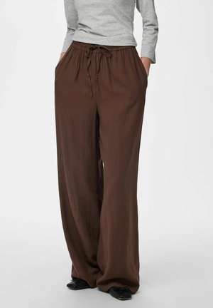 Femme portant un pantalon large marron ample avec taille à cordon, un haut gris à manches longues et des chaussures plates noires, debout les mains dans les poches.