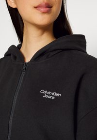 Černá zipová mikina vyrobená z měkké bavlny, která má přední zip a bílé logo "Calvin Klein Jeans" na levé části hrudi.