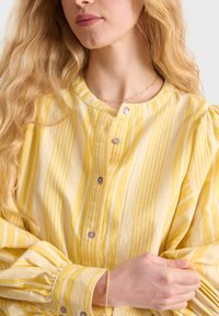Blusa gialla a strisce con chiusura a bottoni, scollatura rotonda e maniche a sbuffo. Realizzata in tessuto morbido, con strisce verticali bianche.