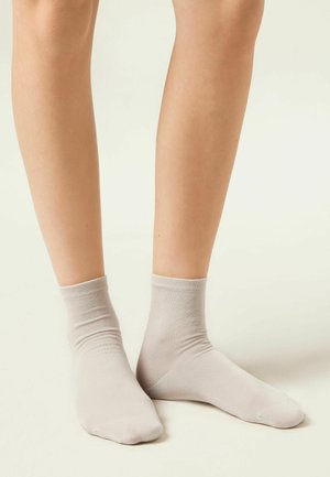 Piernas con calcetines tobilleros gris claro, de pie sobre un fondo claro neutro.