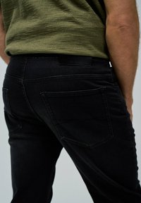 Jeans de mezclilla negra con una camisa texturizada en gris verdoso. Presenta dos bolsillos traseros y diseños de costura sutiles. Silueta minimalista y ajustada.