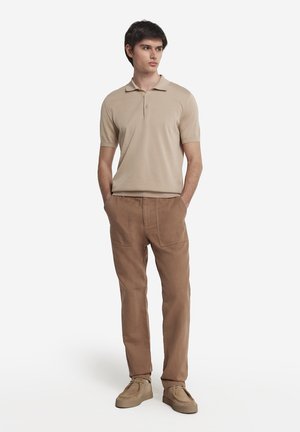 Beige polo shirt med korte ærmer, to-knaps lukning; parret med brune afslappede bukser og lysebrune slip-on sko.