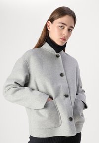 Grå uldjakke med høj krave, der har fire knapper, sidelommer og en afslappet pasform. Teksturen ser blød og glat ud.