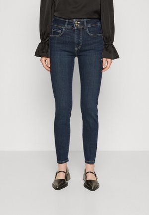 Obcisłe ciemnoniebieskie dżinsy noszone z czarną bluzką z długim rękawem i czarnymi butami na obcasie typu Mary Jane, widoczne od talii do stóp.