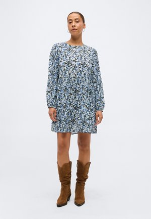 Woman standing wearing a blue floral long-sleeve dress with a round neckline and brown knee-high suede boots.