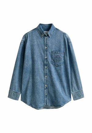 Chemise en denim bleu à manches longues avec boutons-pression, une poche poitrine ornée d'un logo circulaire brodé, et un col à boutons.