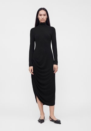 Femme portant une robe midi noire ajustée à manches longues avec col roulé, détails froncés et des ballerines noires pointues avec des accents de boucle.