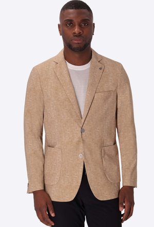Uomo che indossa un blazer beige strutturato sopra una t-shirt bianca e pantaloni neri, in piedi davanti a uno sfondo chiaro e semplice.
