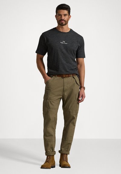 Τιρκουάζ κοντομάνικο μπλουζάκι σε σκούρο γκρι χρώμα με λογότυπο, καφέ παντελόνι cargo με πλευρικές τσέπες και καφέ δερμάτινα μποτάκια με κορδόνια. Χαλαρή εφαρμογή, γυρισμένοι γιακάδες στα παντελόνια.