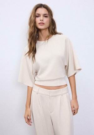 Femme aux cheveux longs et ondulés portant un haut ample beige à manches mi-longues et un pantalon taille haute assorti, sur un fond uni.