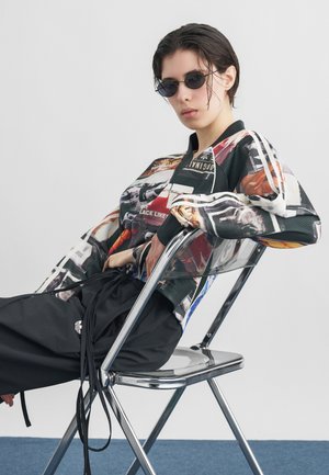 Noor lühikeste märgade juustega, kellel on väiksed ümmargused päikeseprillid ja värvikas graafiline jakk, istub vabal kujul metalltoolil.