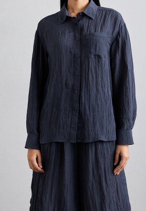 Chemise boutonnée bleu foncé avec une finition texturée, à manches longues, poignets à bouton et une poche poitrine unique. Portée avec une jupe assortie.
