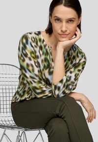 Leichte, gemusterte Bluse in Grüntönen, Blau und Beige mit einem Henley-Ausschnitt und drei Viertel Ärmeln, kombiniert mit dunkelgrünen Hosen.