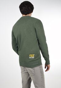 Pull vert à manches longues, avec poignets et ourlet côtelés, arborant un petit logo jaune au dos. Porté avec un pantalon gris clair.