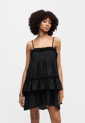 Γυναίκα με σγουρά μαλλιά που φοράει μαύρο αμάνικο φόρεμα με πιέτες και λεπτές τιράντες, στέκεται μπροστά σε ένα απλό φόντο.