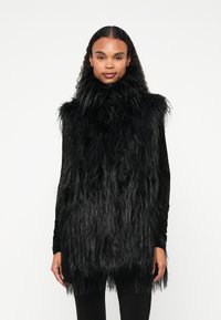 Colete sem mangas em pelúcia preta, com colarinho alto, apresentando um design longo e texturizado, com a bainha ligeiramente ajustada. Forro interior liso visível.