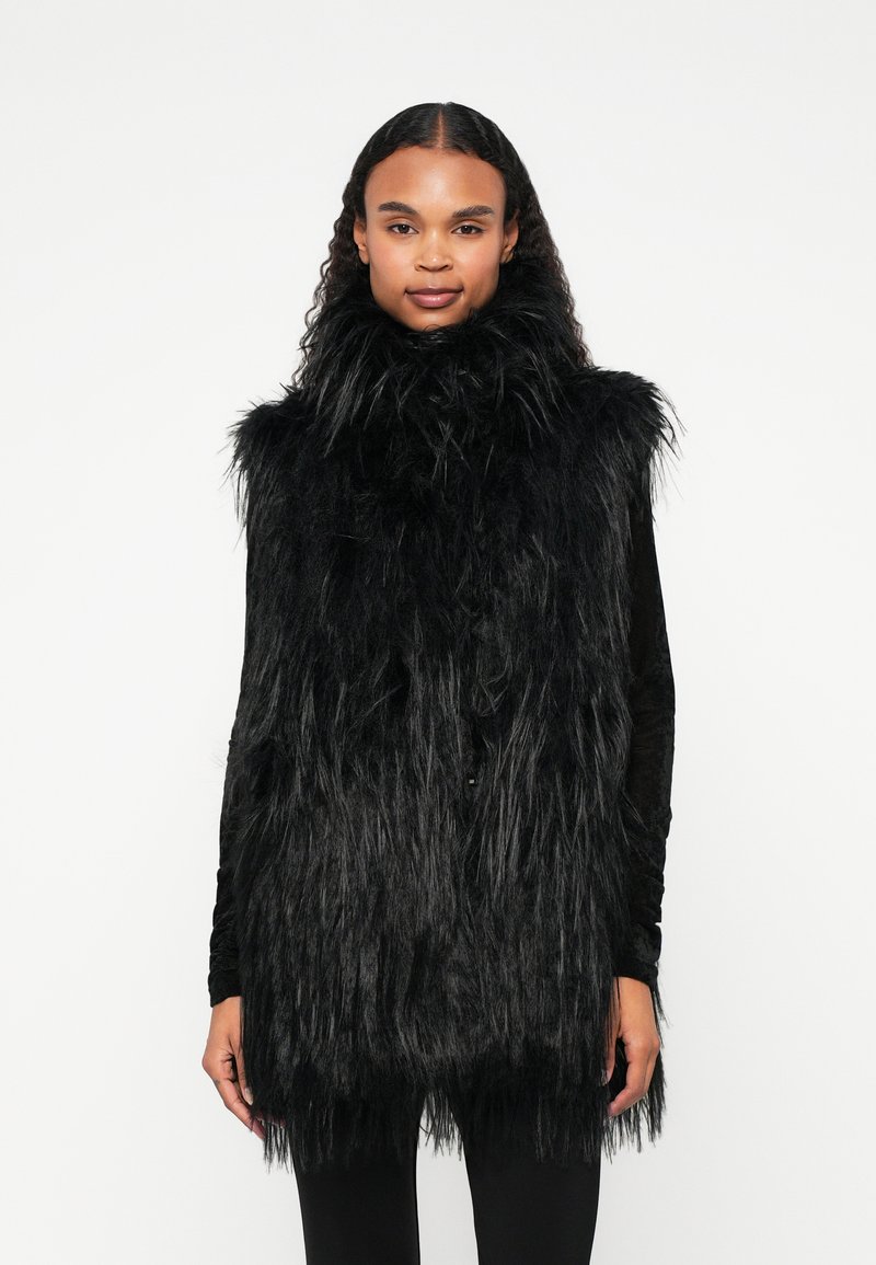 Colete sem mangas em pelúcia preta, com colarinho alto, apresentando um design longo e texturizado, com a bainha ligeiramente ajustada. Forro interior liso visível.