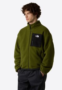 Chaqueta de forro polar verde con un bolsillo en el pecho con cremallera negra, cuello alto y puños ajustables. Presenta un logo en el bolsillo y cremalleras laterales.
