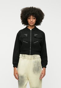 Črna skrajšana bomberska jakna iz mehkega blaga z dvema žepoma na prsih, srebrnimi žebljički na žepih in rebrastimi manšetami.