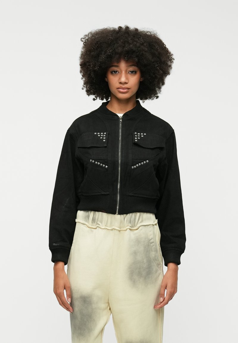 Črna skrajšana bomberska jakna iz mehkega blaga z dvema žepoma na prsih, srebrnimi žebljički na žepih in rebrastimi manšetami.