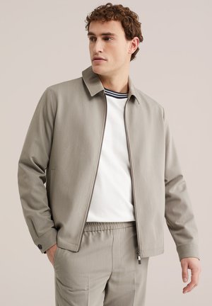 Muškarac obučen u bež jaknu na patentni zatvarač, bijelu košulju s prugastim ovratnikom i bež hlače, stoji ispred jednobojne pozadine i gleda ulijevo.