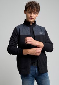 Giacca zip-up nera e blu navy con colletto alto, pannello superiore a contrasto e polsini a costine. Presenta tasche laterali e logo "BIG STAR".
