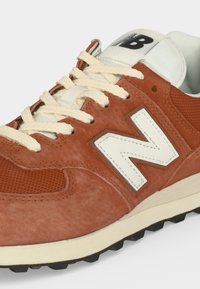 Καφέ σουέντ αθλητικό παπούτσι New Balance με λευκό λογότυπο "N", εκρού κορδόνια, λευκή γλώσσα με μαύρο "B" και υφασμάτινο καφέ πλέγμα κοντά στα δάχτυλα.