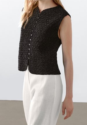 Haut noir texturé sans manches à boutons associé à un pantalon blanc ample taille haute sur une personne debout.