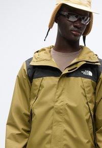 Απωθητικό νερού κίτρινο-πράσινο μπουφάν με μαύρες λεπτομέρειες, εμπρόσθιο φερμουάρ και ρυθμιζόμενο κουκούλα. Το υλικό φαίνεται λείο και ελαφρύ.